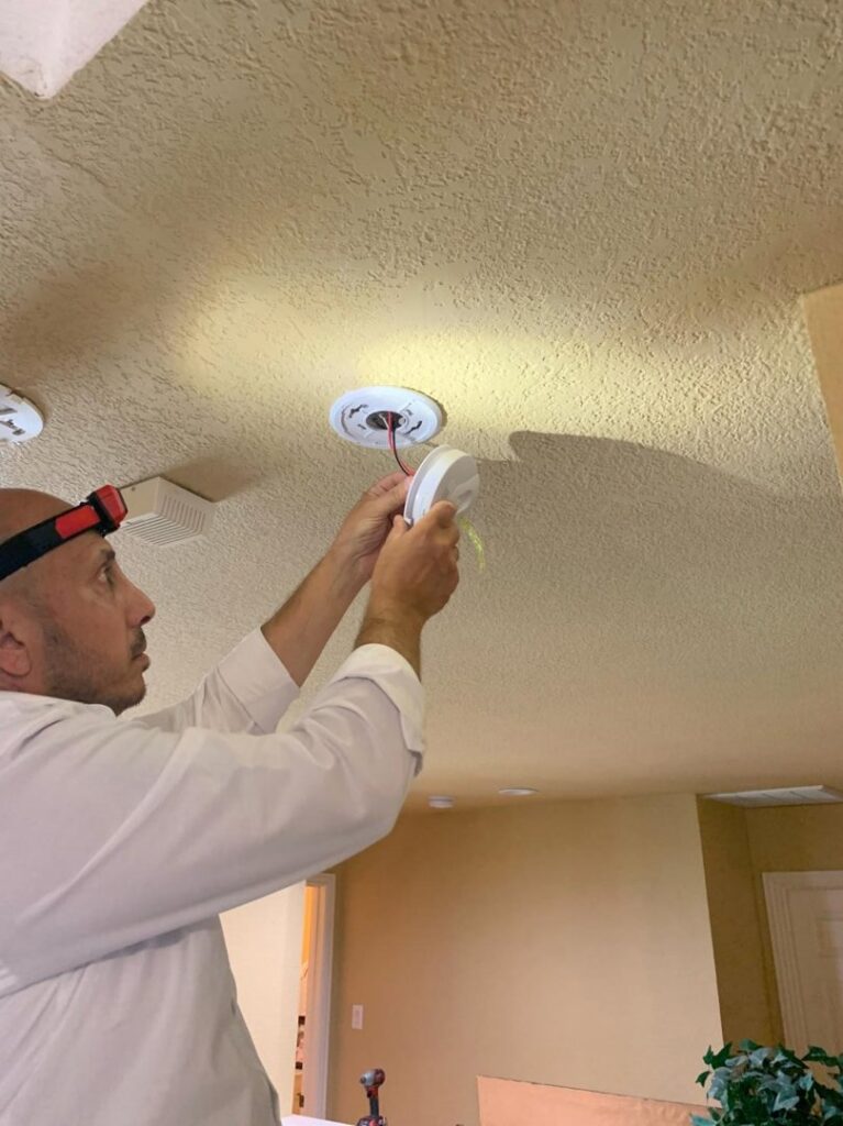 An electrician wearing a headlamp installing a ceiling smoke detector or recessed light fixture, a service from Volta Electrical LLC in Plano, TX.