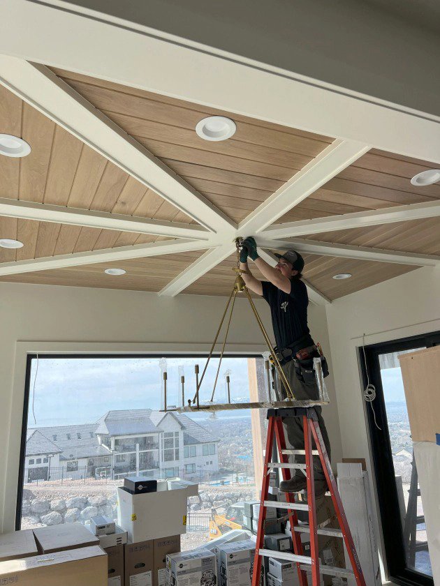An electrician from Homestead Electric on a ladder installing a chandelier in a ceiling with recessed lights in Orem, UT.