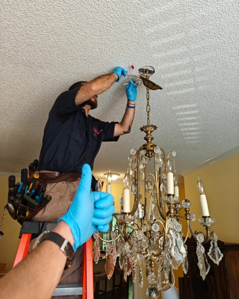 An electrician on a ladder installing a decorative chandelier, with a thumbs-up, for Sherrill Company, LLC Electrical Contractors in Columbus, GA.