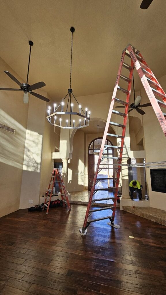 An electrician from V Electrical LLC installing a new chandelier in a spacious living room in Albuquerque, NM.