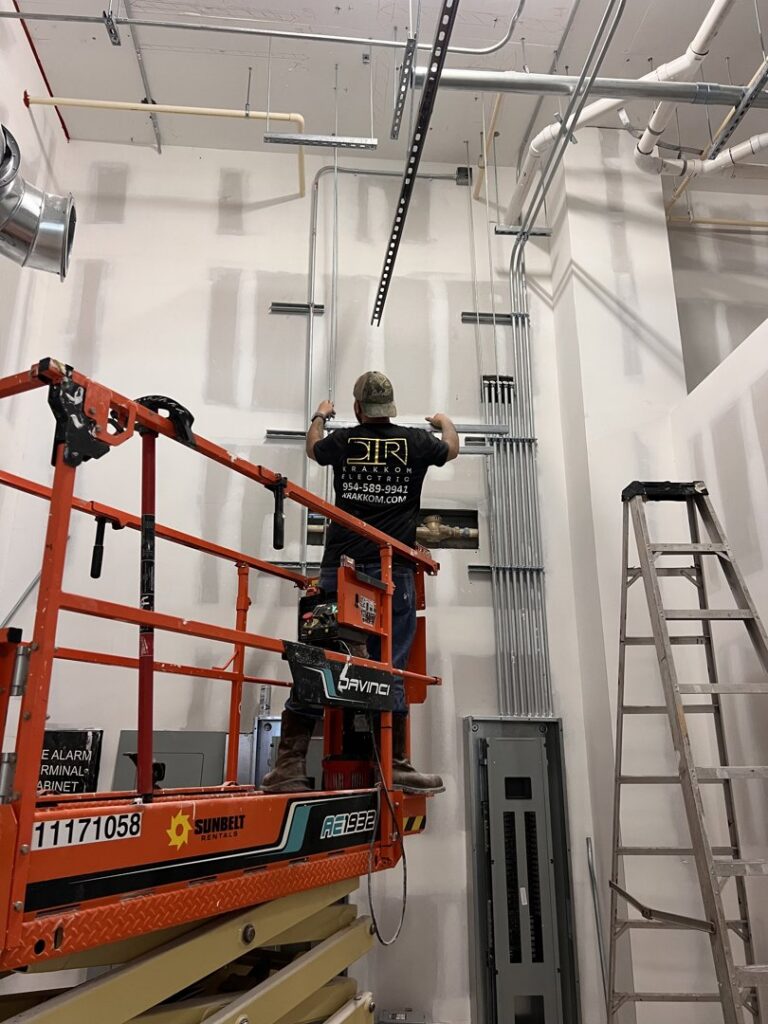 An electrician on a scissor lift installing electrical conduit and panels in a commercial building for Krakkom Electric in Sunrise, FL.