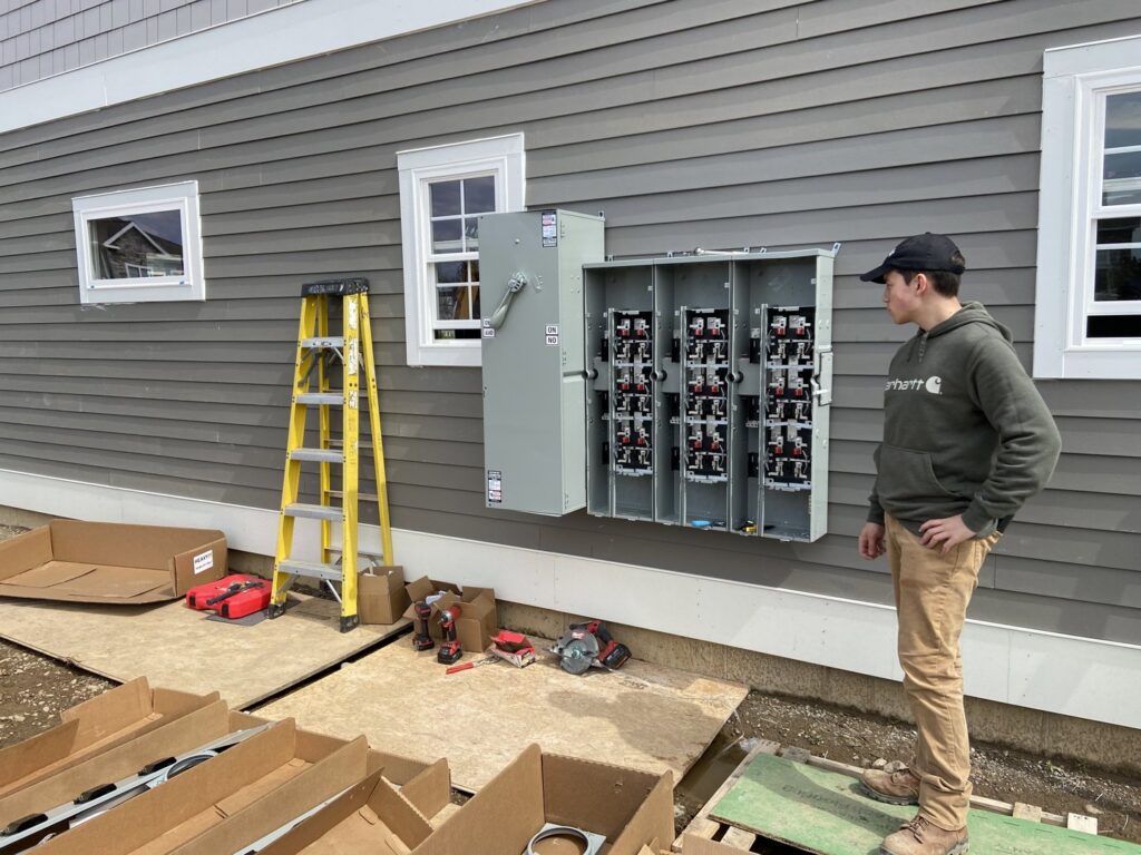 An electrician standing next to an open electrical panel being installed on the exterior of a new construction home by Greif Electric Company in Dolgeville, NY.