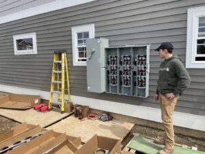 An electrician standing next to an open electrical panel being installed on the exterior of a new construction home by Greif Electric Company in Dolgeville, NY.