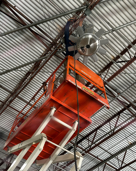 An electrician installing a large industrial fan and light fixture from a scissor lift for Rubio Electrical Innovations LLC in San Antonio, TX.