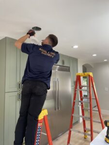 An electrician on a ladder installing a recessed light fixture in a modern kitchen for Miller and Sons Electric in North Myrtle Beach, SC.