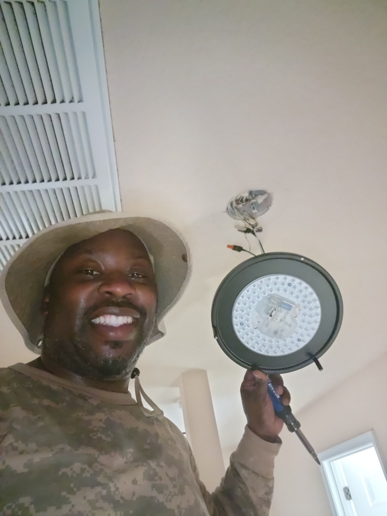 An electrician from Rain Man Thee Handyman installing a new LED light fixture in Hampton, VA.