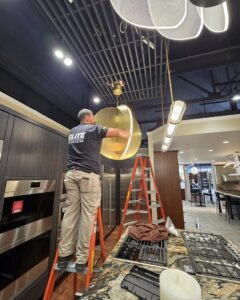 An Elite Electric Company electrician on a ladder installing a large, modern light fixture in a kitchen showroom in Melrose Park, IL