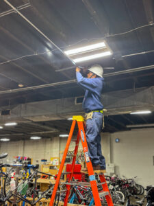 An electrician on a ladder installing or repairing an overhead light fixture, a service provided by Carolina Voltage in Charlotte, NC.