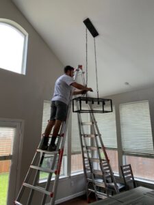 An electrician on a ladder installing a large, modern chandelier in a high-ceiling room, a service from Volta Electrical LLC in Plano, TX.