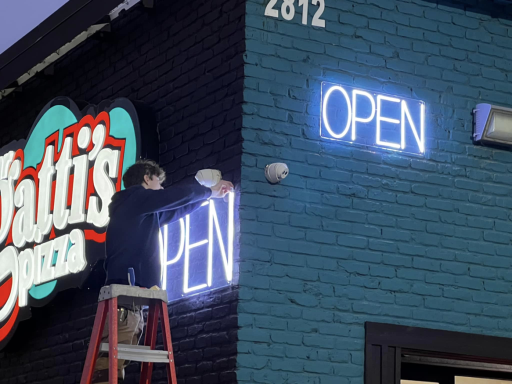 An electrician on a ladder installing a neon 'OPEN' sign on a commercial building for APEX Electrical Design in Fort Worth, TX