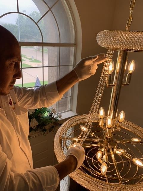 An electrician wearing gloves carefully installing an ornate chandelier, a service provided by Volta Electrical LLC in Plano, TX.