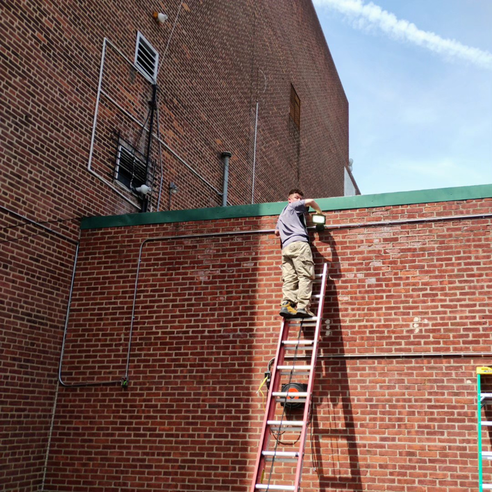 An electrician installing an outdoor light fixture on a brick building for C&J And Sons Multi Services in Newark, NJ.