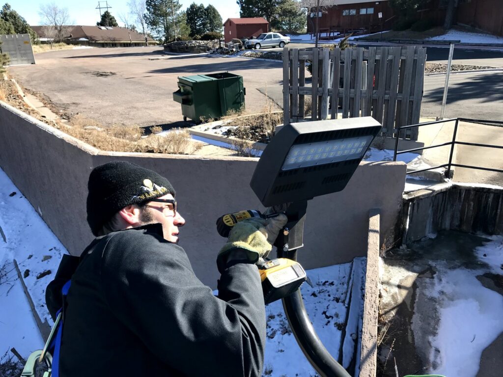 An electrician from The Electric Way in Colorado Springs, CO, installing an outdoor light fixture with a drill.