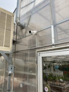 An electrician from D.B. Smart Homes & Security installing a security camera and electrical box on a greenhouse in Sheridan, WY.