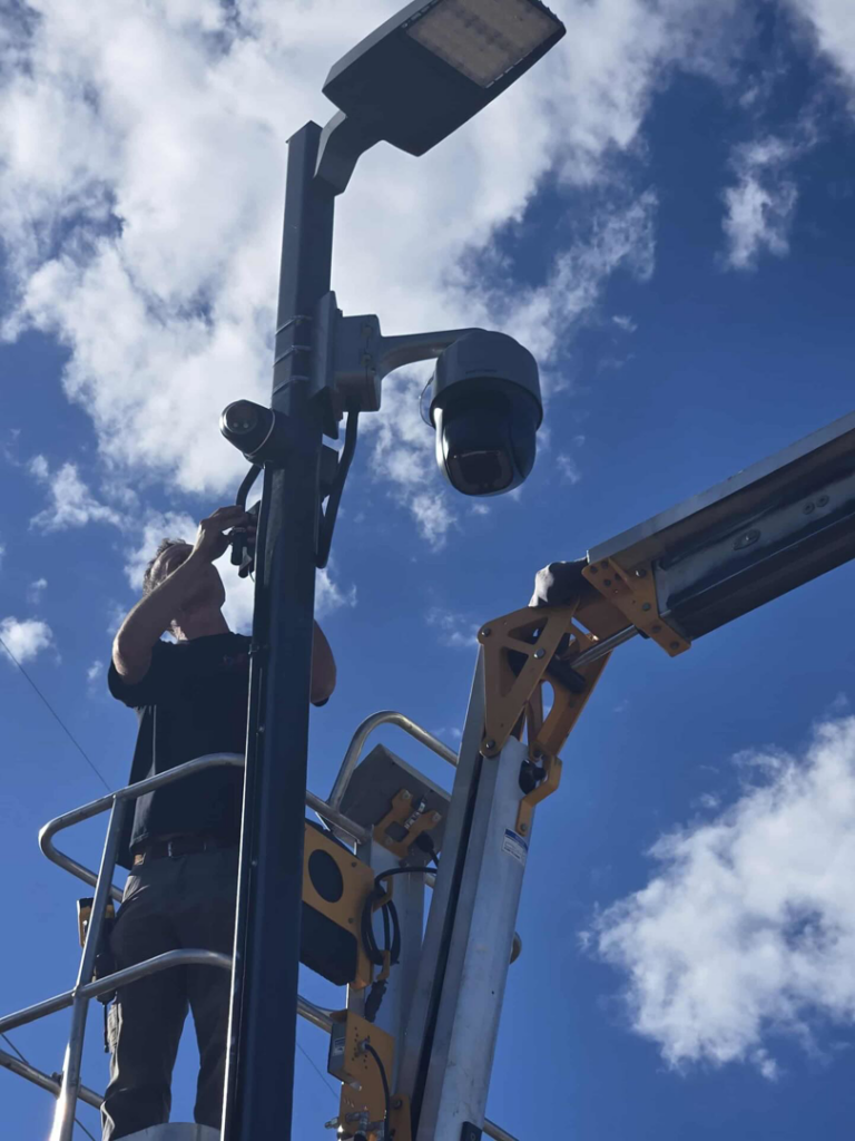 An electrician from D.B. Smart Homes & Security LLC installing a street light and security camera on a pole in Sheridan, WY.
