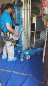 An electrician moving a portable lift, wearing shoe covers and a tool belt, for Peace Of Mind Services in Sag Harbor, NY