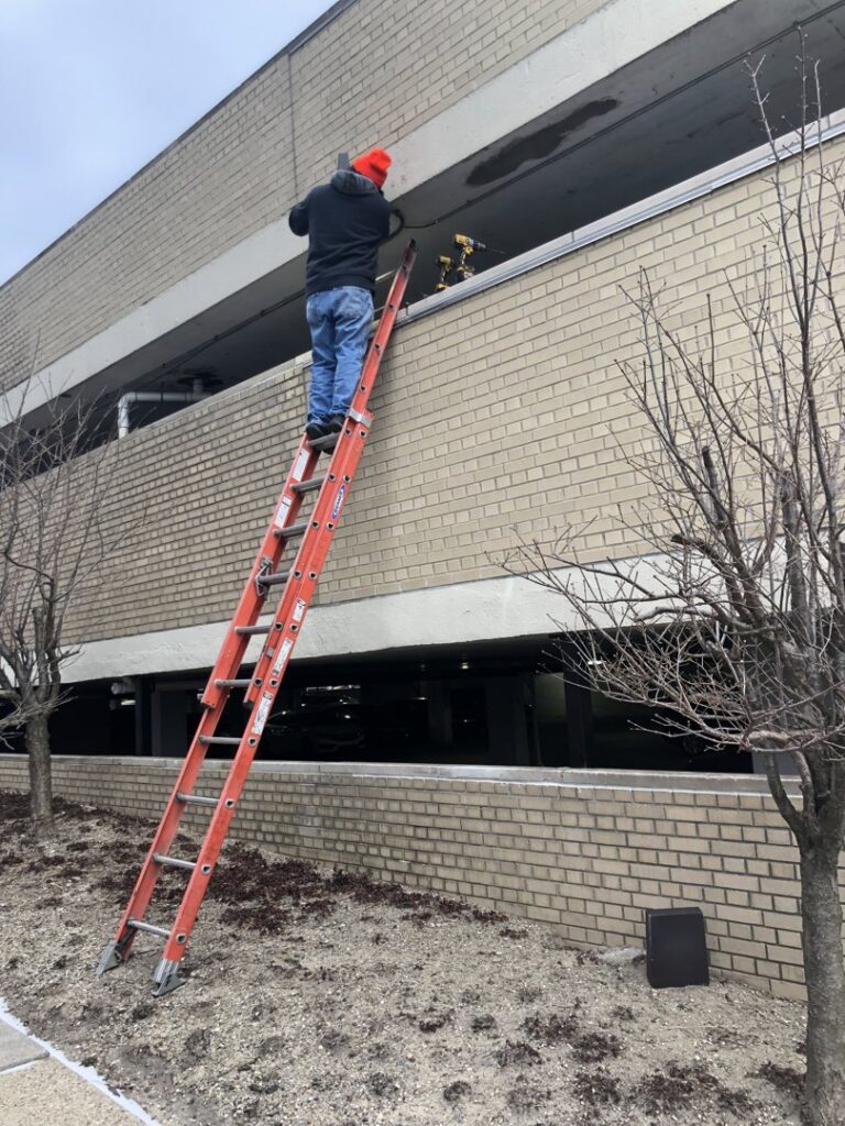 An electrician on a ladder installing or repairing exterior lighting for Detroit Voltage in Detroit, MI.