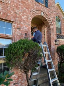 An electrician on a ladder working on exterior wiring or a light fixture near the front door of a brick house by Volta Electrical LLC in Plano, TX.