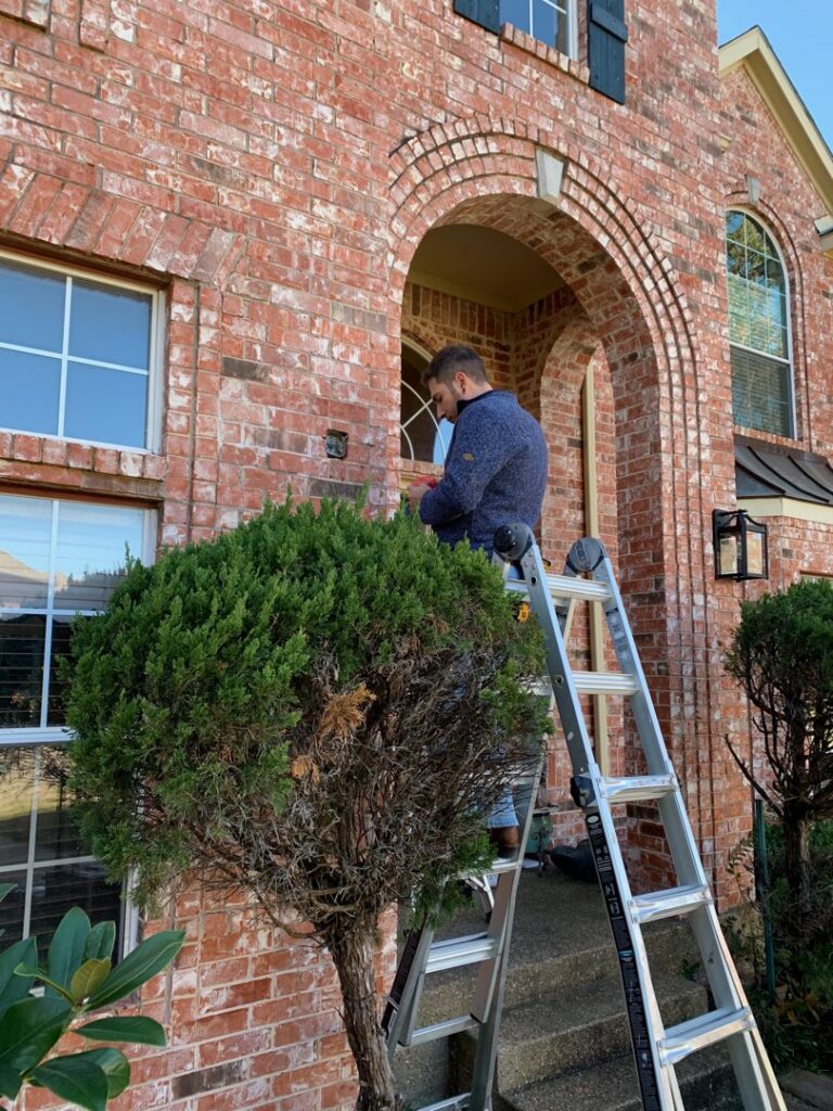 An electrician on a ladder working on exterior wiring or a light fixture near the front door of a brick house by Volta Electrical LLC in Plano, TX.