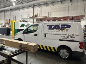 A TAP and Sons Electric service van with a ladder on top, parked inside a warehouse in Tempe, AZ.