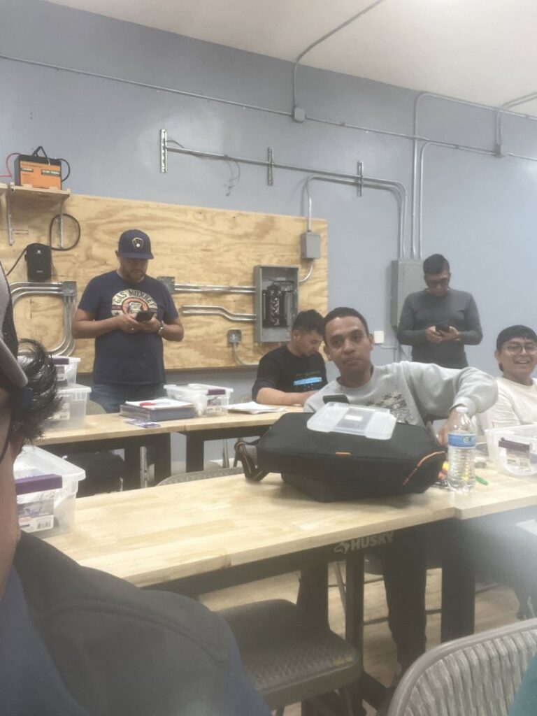 Students in an electrician training classroom with electrical panels and conduit visible on the wall at Escuela Tecnica ABC in North Chicago, IL