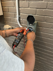 An electrician testing an outdoor electrical outlet with a clamp meter, ensuring safety and functionality for AHM ELECTRIC in Houston, TX.