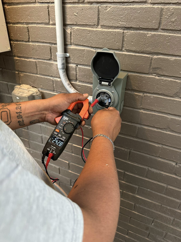 An electrician testing an outdoor electrical outlet with a clamp meter, ensuring safety and functionality for AHM ELECTRIC in Houston, TX.