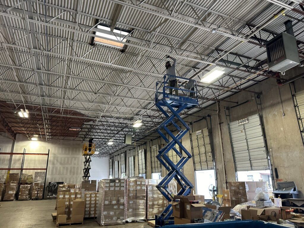 An electrician on a scissor lift installing or maintaining overhead lighting in a warehouse for Rubio Electrical Innovations LLC in San Antonio, TX.