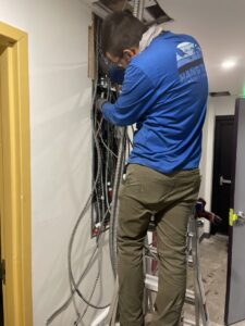 An electrician on a ladder working on complex electrical wiring and conduits inside a wall for Flowers Electric in Aurora, CO.