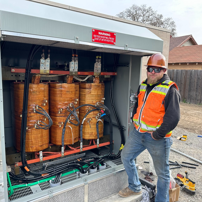 An electrician in safety gear stands next to a large electrical transformer at Power City Electric in Spokane, WA.
