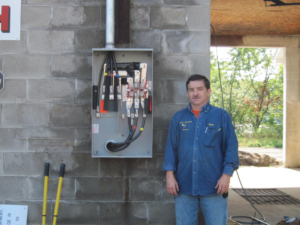 An electrician from Dean's Electrical Service standing next to an open electrical service panel in Corinth, NY.