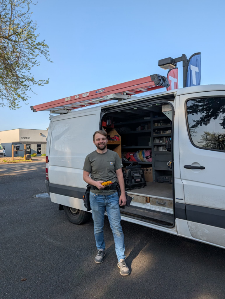 An electrician with a tool belt and voltage tester standing by a service van for Zoom Electric in Vancouver, WA.