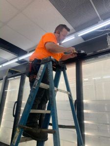 An electrician on a ladder working on wiring above commercial refrigeration units for Lightningwerks Electrical in Springfield, MO.