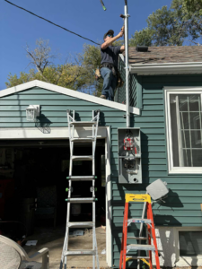 An electrician working on an electrical mast and service panel on a house in Minot, ND, by Black Electric & Sound.