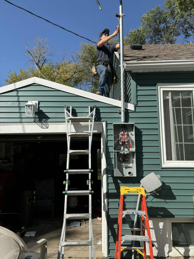 An electrician working on an electrical mast and service panel on a house in Minot, ND, by Black Electric & Sound.