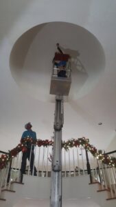 An electrician on a lift working on a high ceiling light fixture, observed by a colleague, for Peace Of Mind Services in Sag Harbor, NY