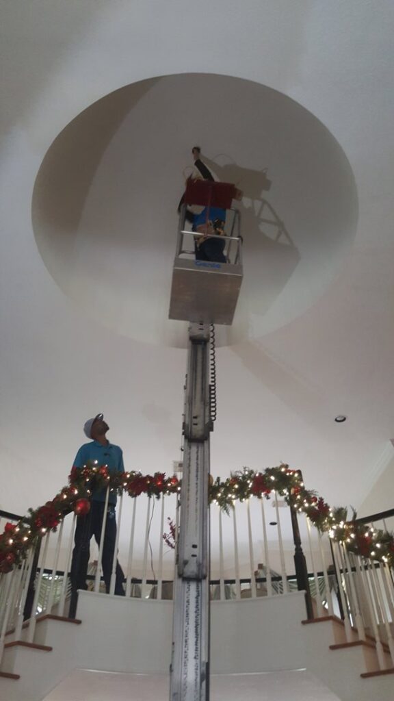 An electrician on a lift working on a high ceiling light fixture, observed by a colleague, for Peace Of Mind Services in Sag Harbor, NY