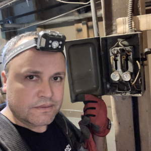 An electrician from Diaz Techician LLC in Chicago, IL, working on an old electrical panel with exposed wiring and fuses.