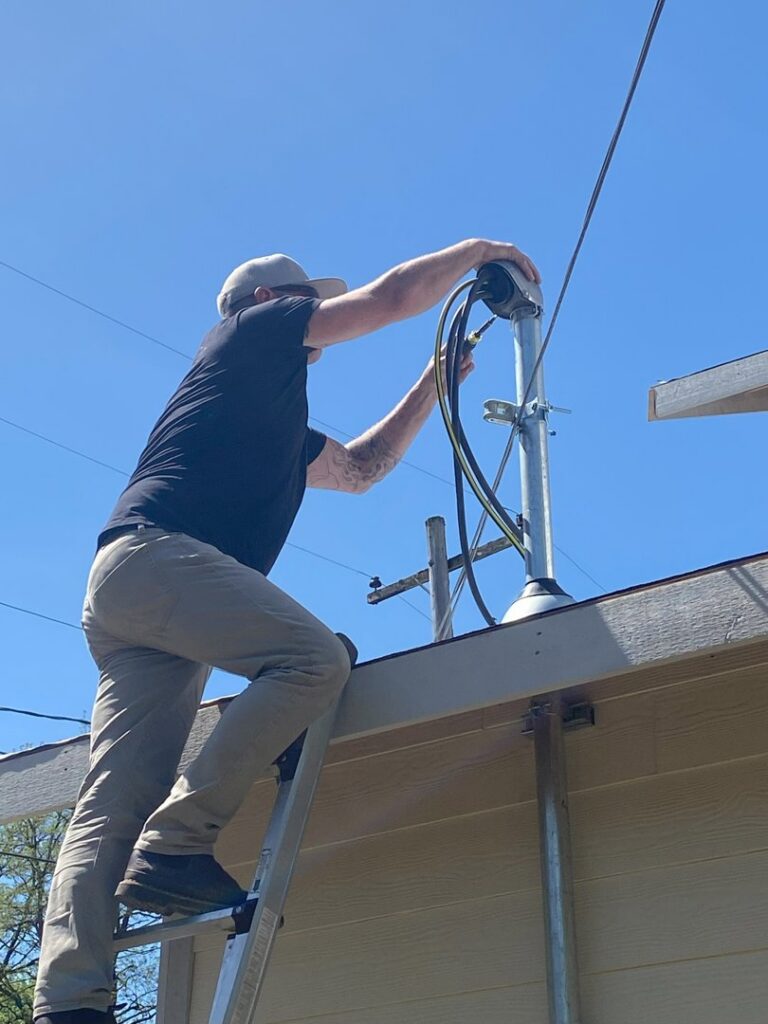 An electrician on a ladder working on an outdoor electrical service mast, performing an installation or repair for Devaney Electric Co. in Lacey, WA.