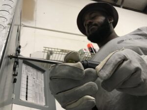 An electrician from AK Electrical, LLC wearing gloves and a company shirt, working on an electrical panel with a screwdriver in Mesquite, TX.