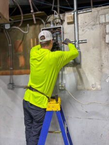 An electrician on a ladder working on an electrical panel, performing service for Carolina Voltage in Charlotte, NC.