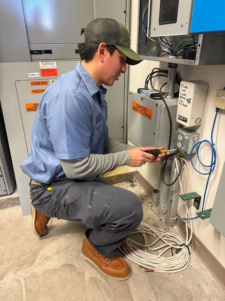 An electrician working on wiring near electrical panels with a "Danger High Voltage" sign at Three Rivers Electric in Fort Wayne, IN.