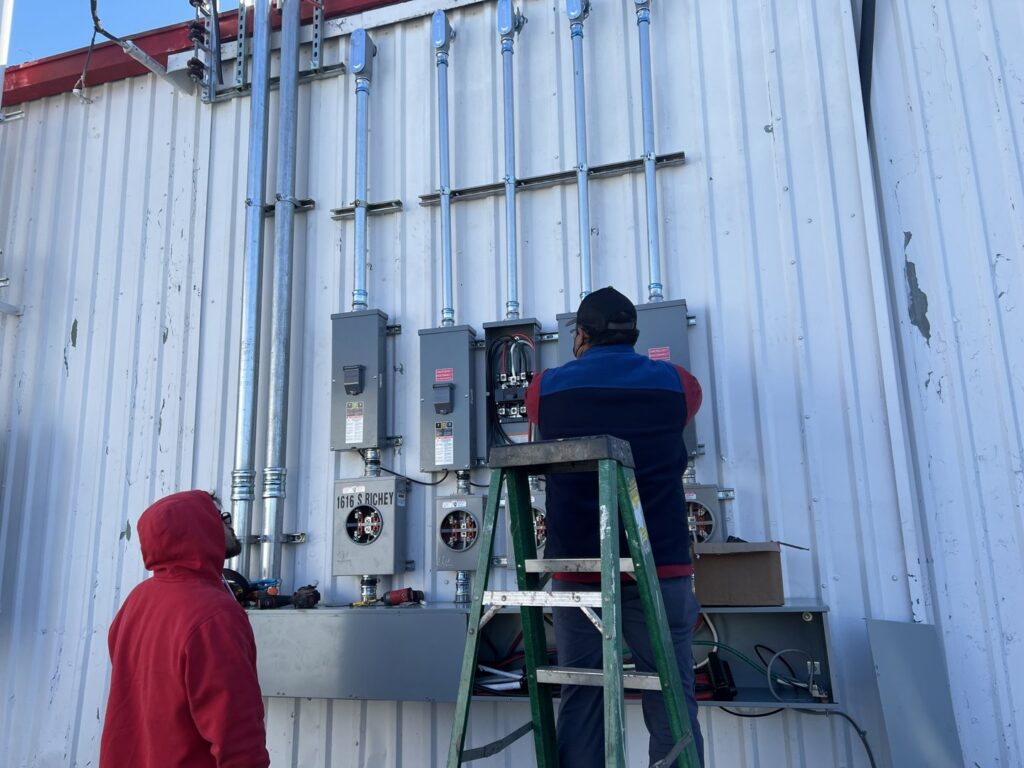 Two electricians installing multiple commercial electrical panels and conduits on an exterior wall for Llanos Electric Builders in Houston, TX.