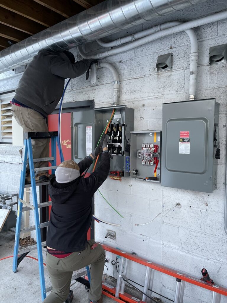 Two electricians installing and wiring electrical panels and conduits on an exterior wall for Flowers Electric in Aurora, CO.