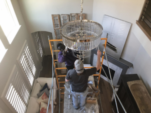 Two electricians on scaffolding installing a large, ornate chandelier in a home, provided by APEX Electrical Design in Fort Worth, TX