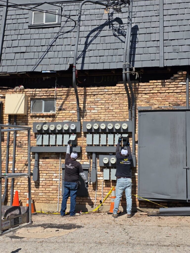 Two electricians from Acevedo's Electric installing outdoor electrical meters and panels on a brick building in Dallas, TX.