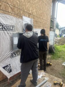 Two electricians from Andrew's Electric installing an outdoor electrical panel on a building in Kent, WA