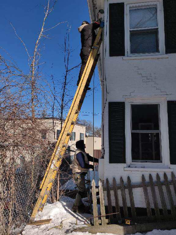 Two electricians from Lindquist Electric on a ladder installing or repairing exterior wiring on a house in Upton, MA.