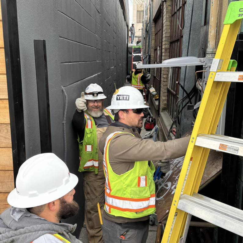 Electricians in hard hats and safety vests working with ladders in a narrow alleyway for Wheeler Electric, Inc. in Idaho Falls, ID.