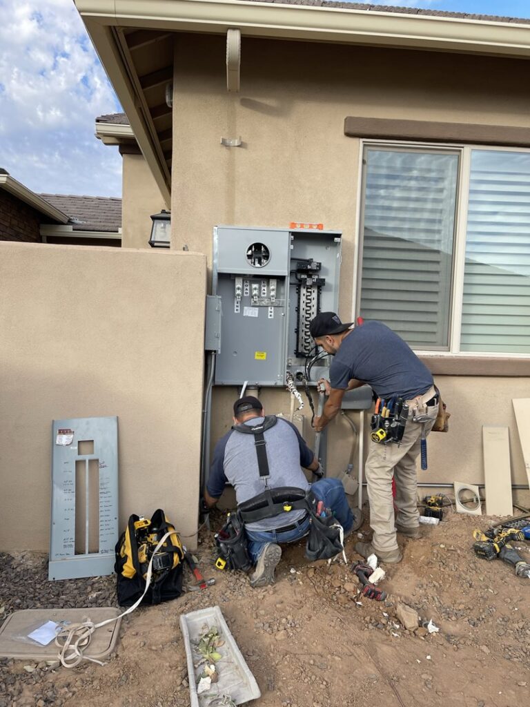 Two electricians from EV Electric LLC working on an exterior electrical panel and meter base in Mesa, AZ